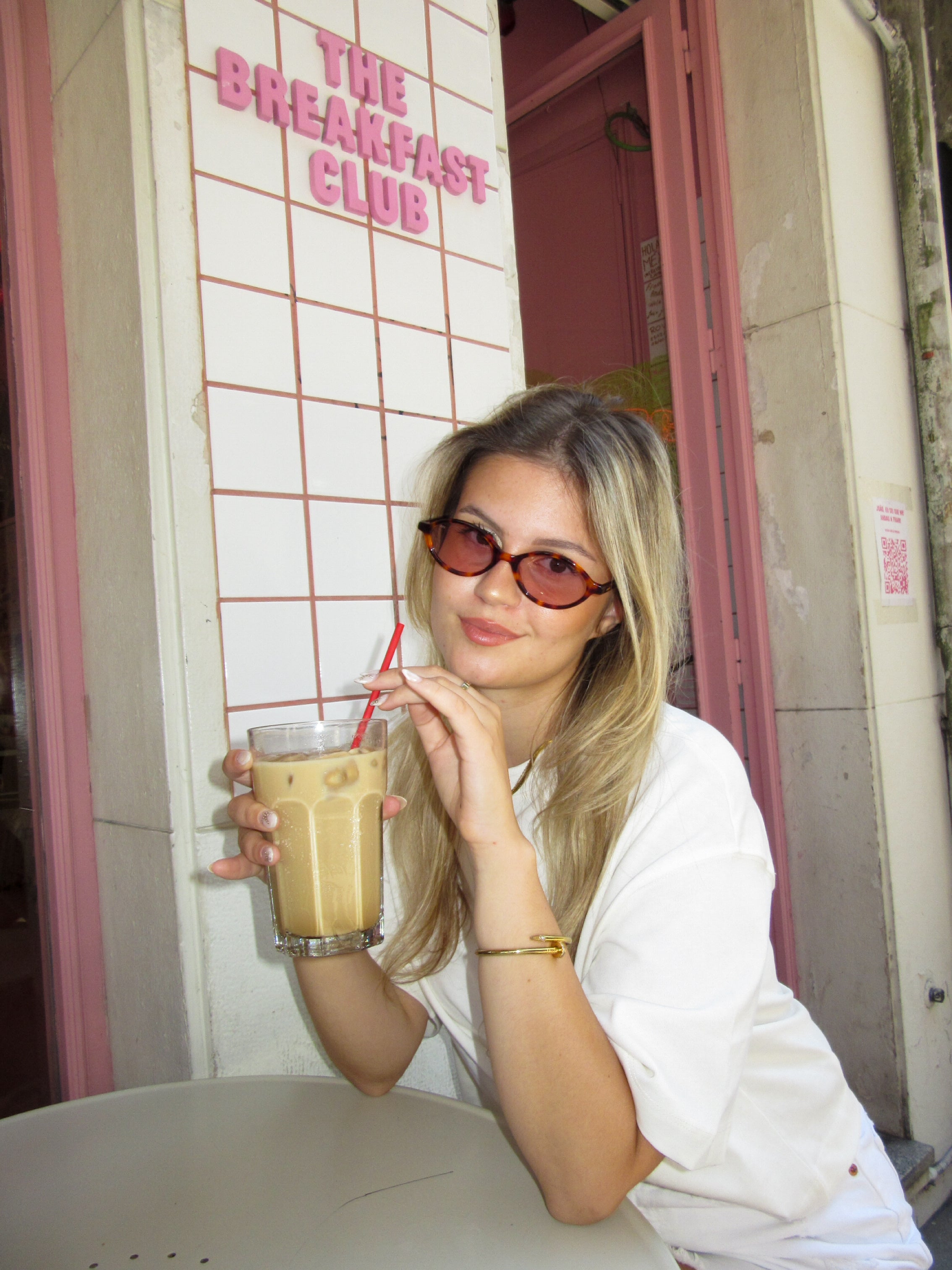 Woman holding a drink with a red straw in front of 'The Breakfast Club' sign. With Misu Sunglasses