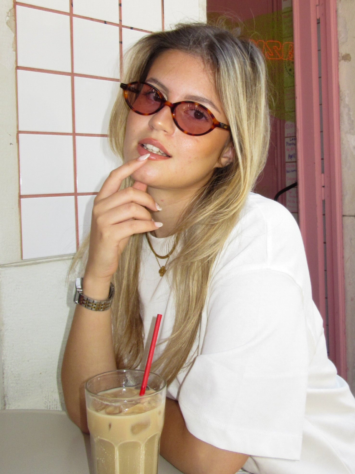 Woman sitting at a table with a drink, wearing Misu Monelise sunglasses and a white shirt, in front of a 'The Breakfast Club' sign.
